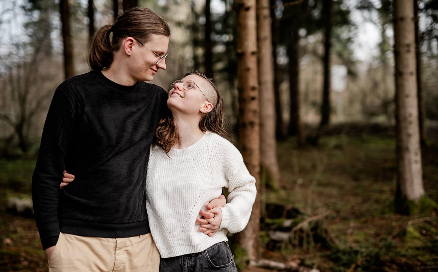 Zwei Personen stehen lächelnd im Wald und sehen einander an.