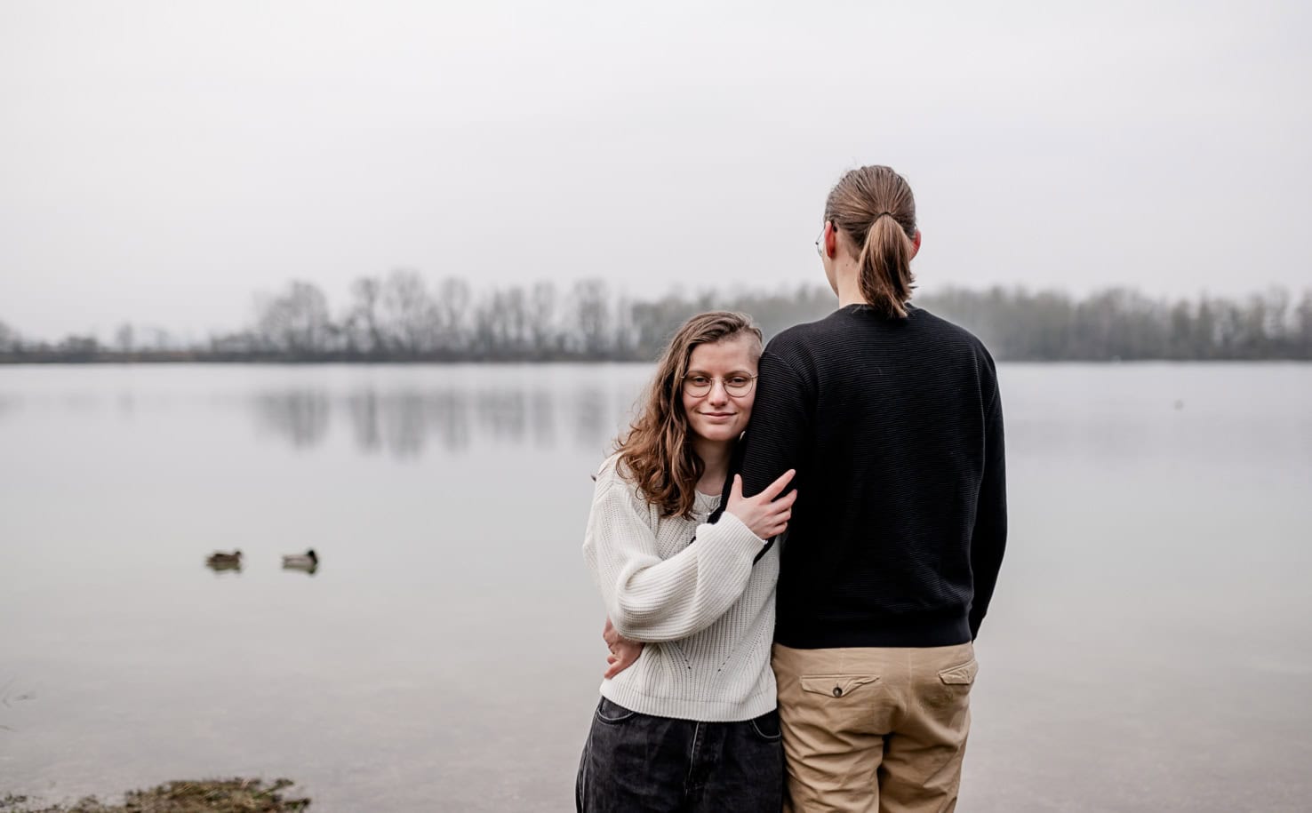 Das Bild zeigt zwei Personen, die sich am Ufer eines Sees aneinander kuscheln, wobei die eine Person in die Kamera blickt.