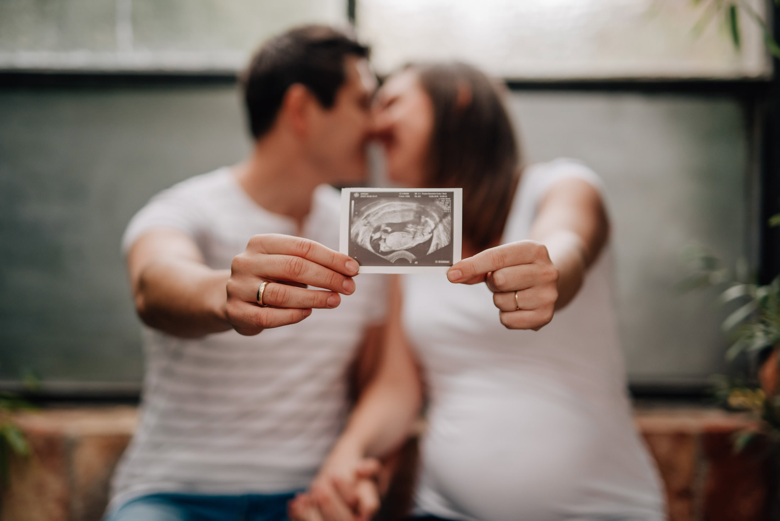 Das Bild zeigt ein küssendes Paar, das gemeinsam ein Ultraschallbild eines ungeborenen Babys hält.