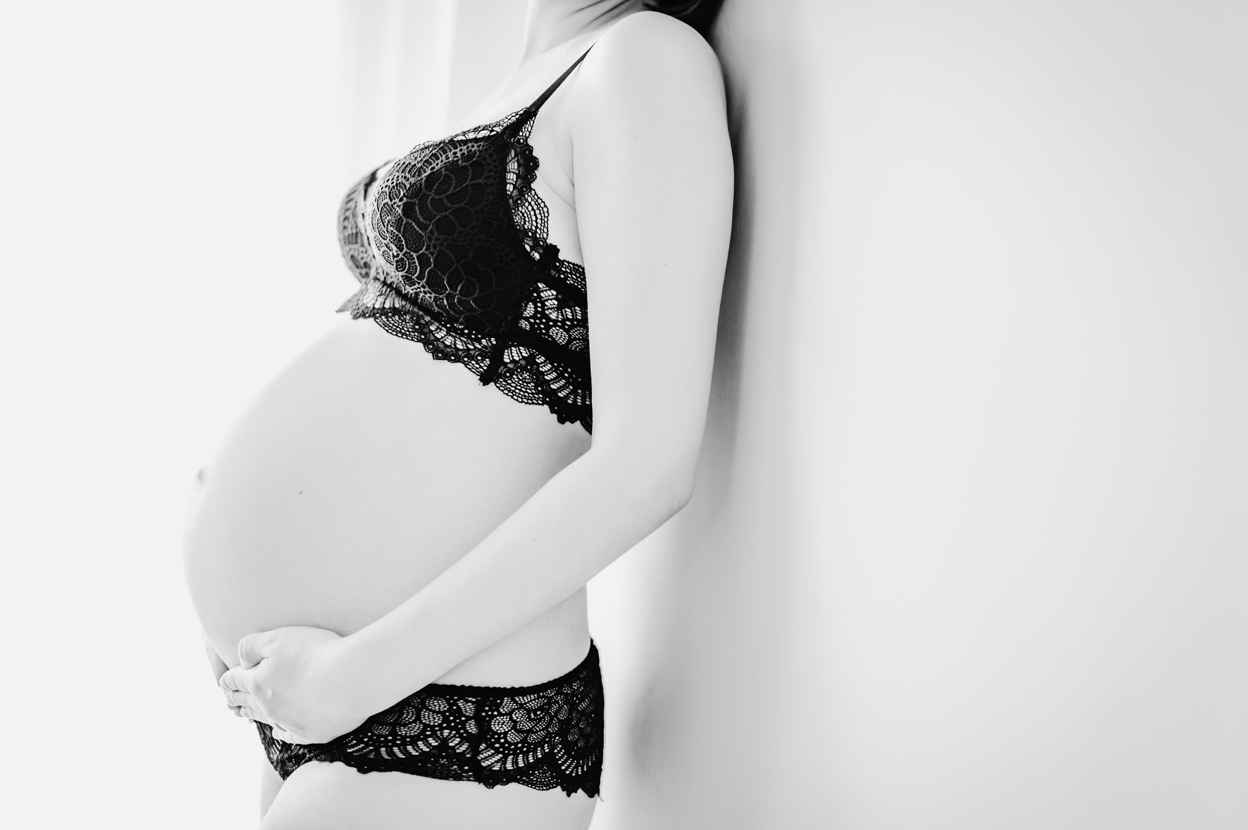 Das Bild zeigt eine schwangere Frau, die schwarze Spitzenunterwäsche trägt und ihren Bauch hält.