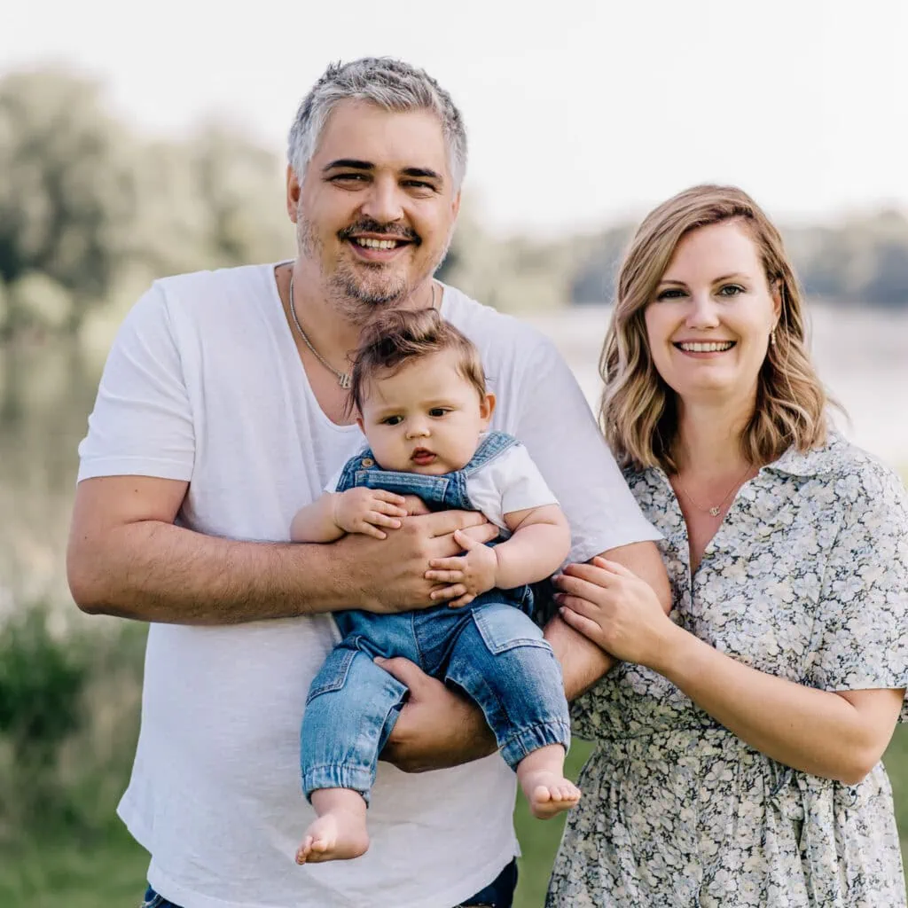 Das Bild zeigt eine Familie mit zwei Erwachsenen und einem Baby, die gemeinsam lächeln.