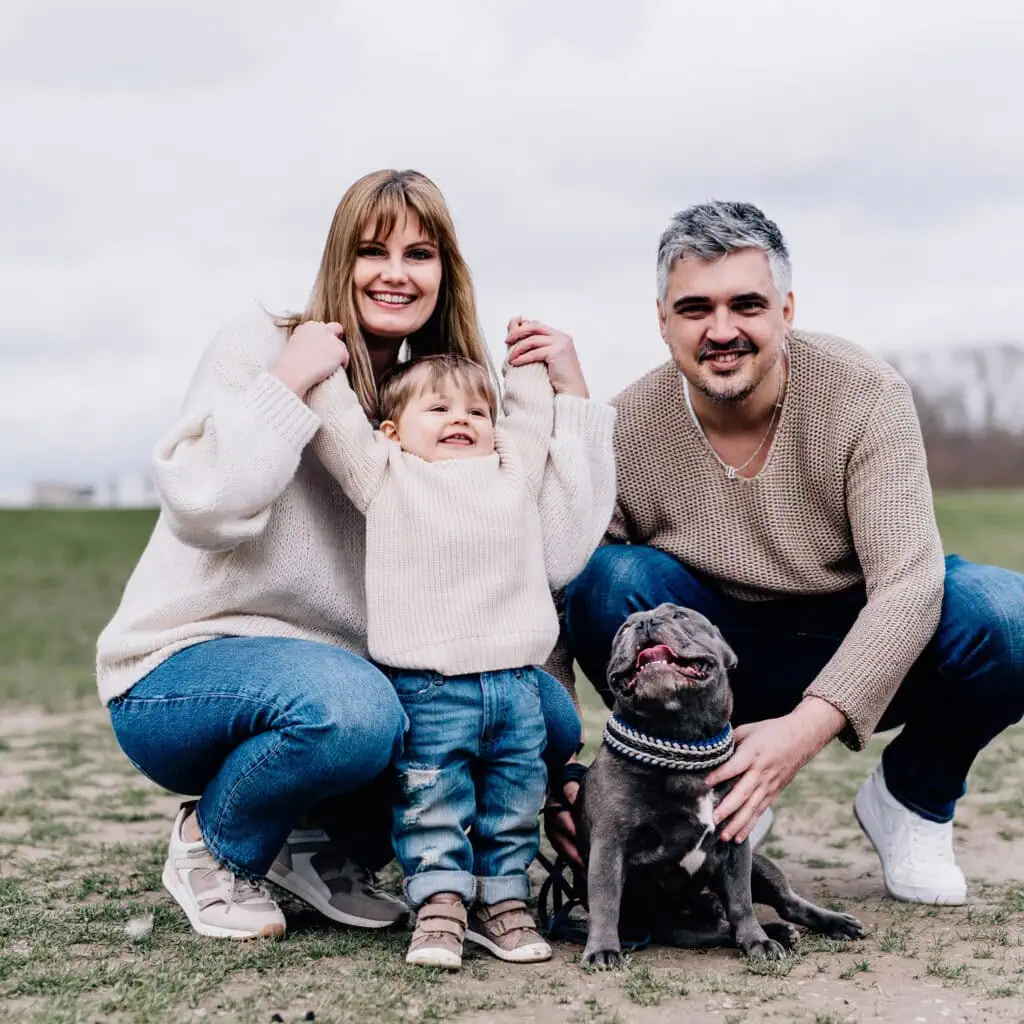 Das ist ein Foto von zwei Erwachsenen und einem Kind, die lächelnd im Freien sitzen, neben ihnen befindet sich ein Hund.