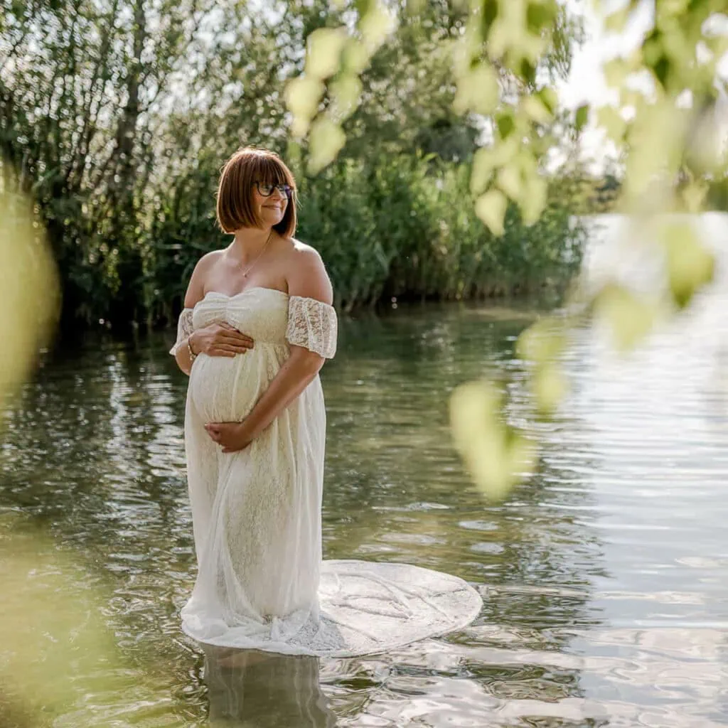 Eine schwangere Frau steht in einem weißen Kleid im Wasser und hält ihren Bauch.