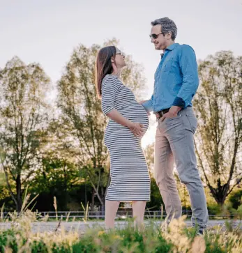 Das Bild zeigt zwei Personen, die sich anlächeln und eine schwangere Frau halten, in einem sonnigen, parkähnlichen Umfeld.