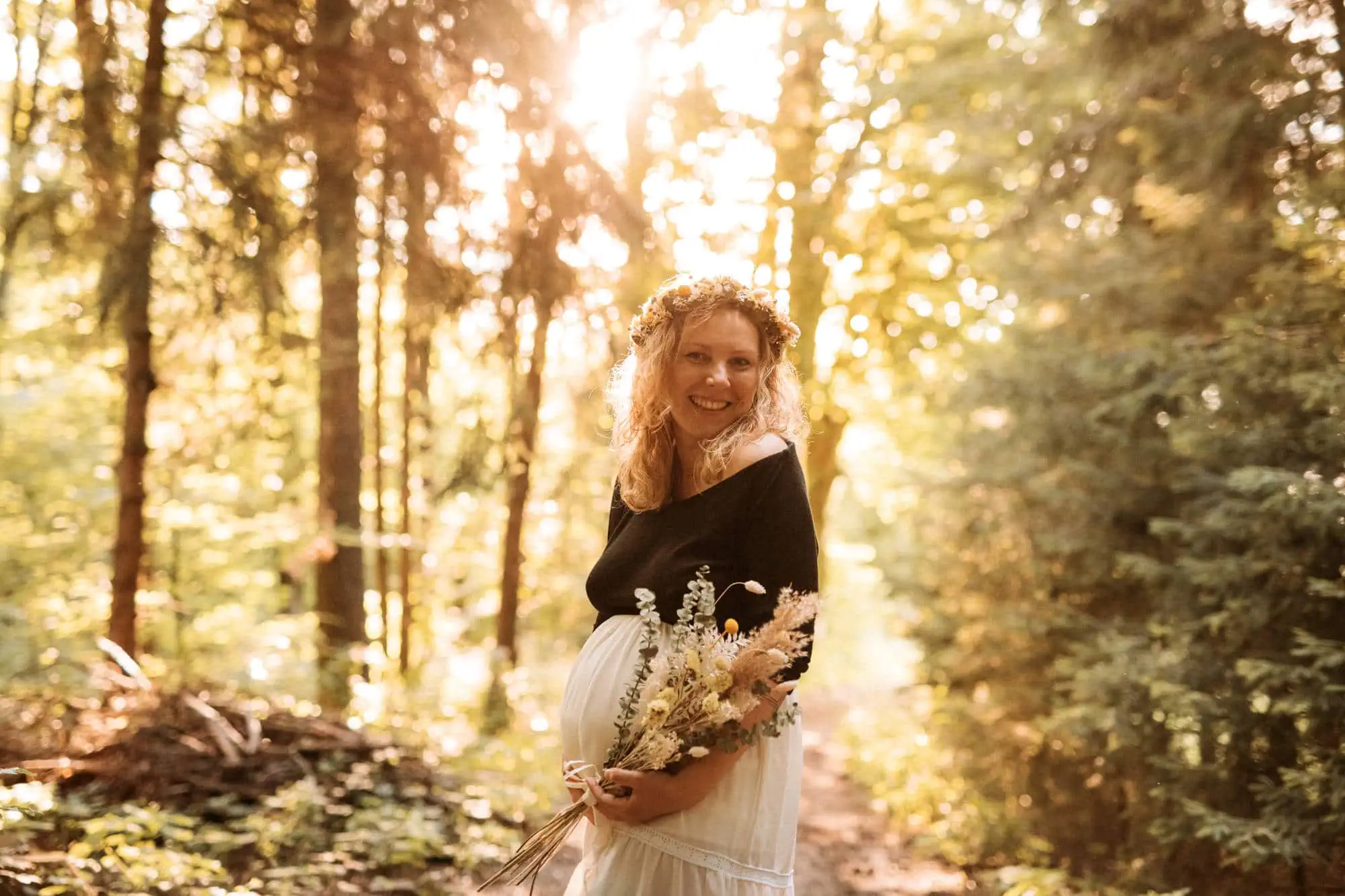 Auf dem Bild ist eine lächelnde Frau mit einem Blumenkranz im Haar, die in einem Wald steht und einen Blumenstrauß hält.