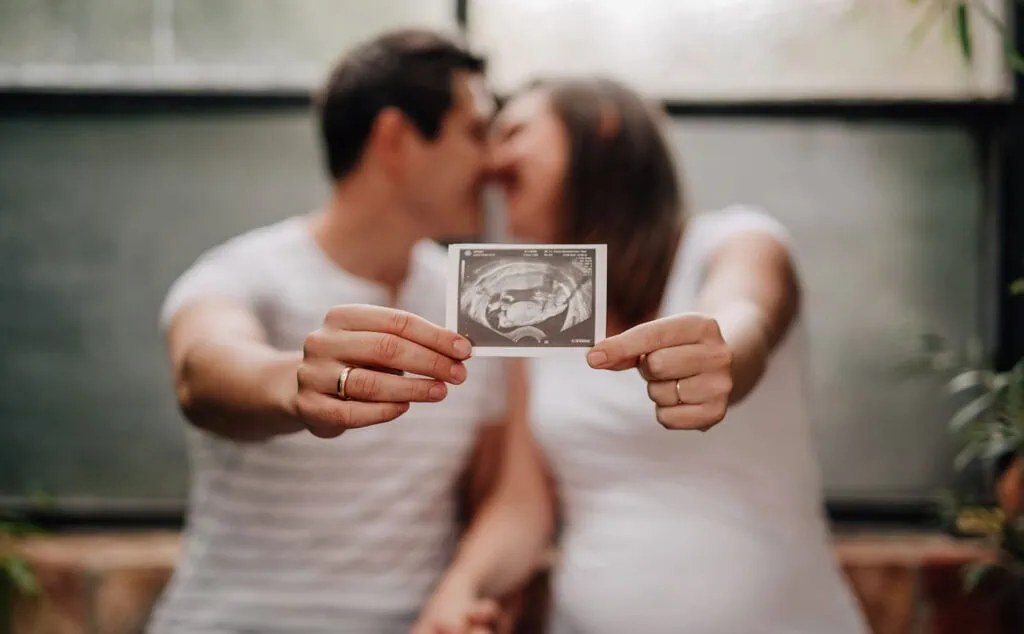 Ein Paar küsst sich im Hintergrund und hält gemeinsam ein Ultraschallbild eines Babys in die Kamera.