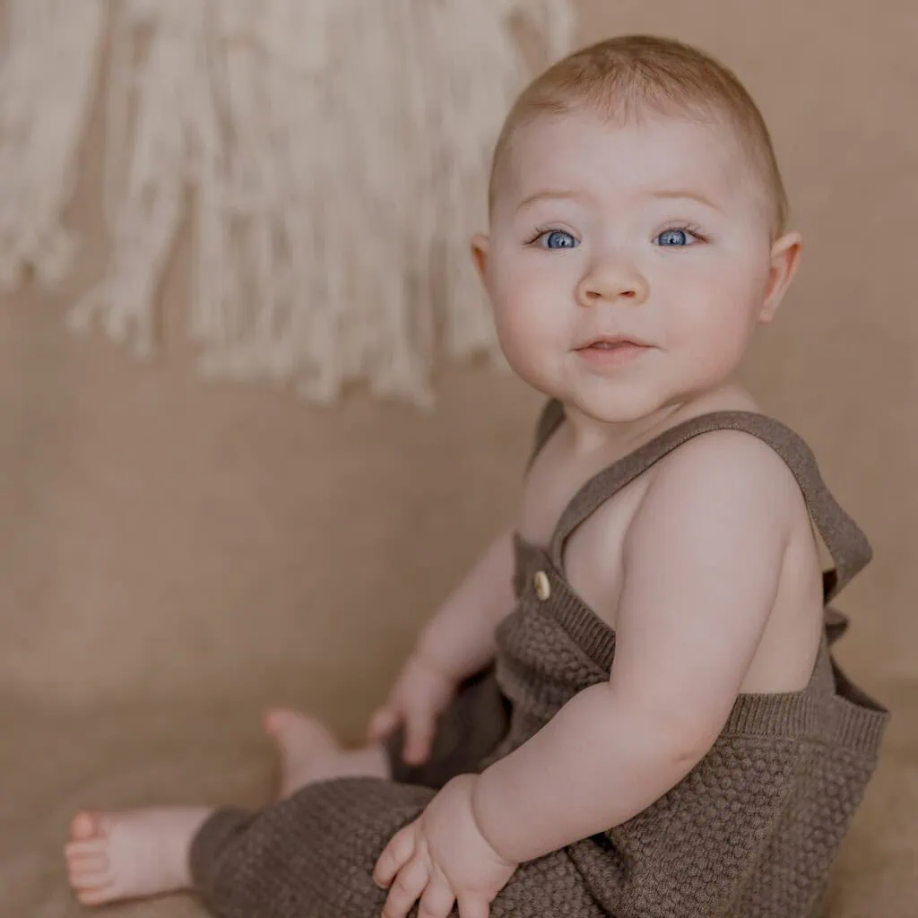 Das ist ein Bild von einem lächelnden Baby in beiger Kleidung, das sitzt.