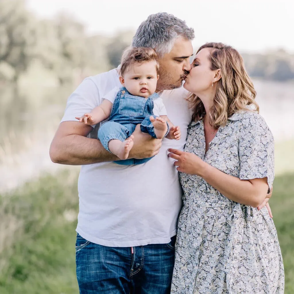 Das Bild zeigt einen Mann und eine Frau, die sich einen Kuss geben, während der Mann ein Baby hält.