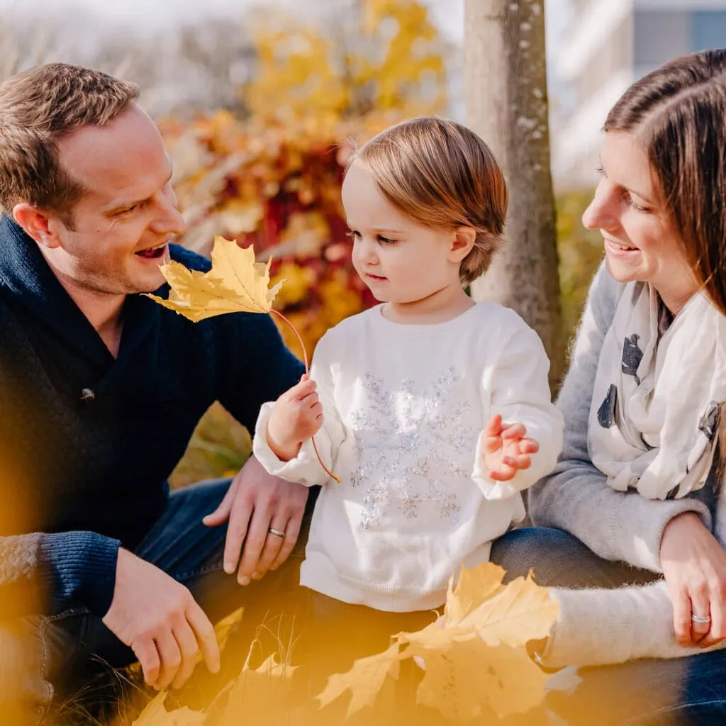 Ein Kind hält Herbstblätter, während es zwischen zwei lächelnden Erwachsenen sitzt.