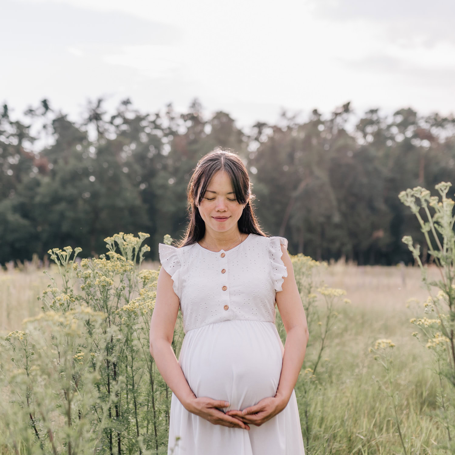 Eine schwangere Frau hält ihre Hände um ihren Bauch und steht auf einer Wiese.