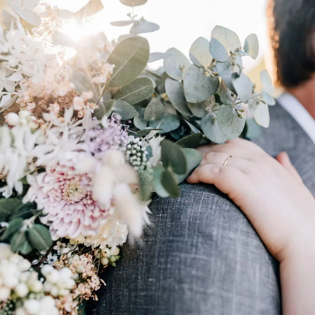 Das Bild zeigt eine Person, die einen blumengeschmückten Arm um eine andere Person legt, wobei eine Hochzeitsring am Finger zu sehen ist.