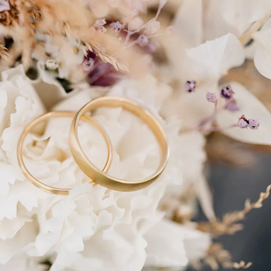 Das Bild zeigt zwei goldene Ringe auf einem floralen Hintergrund, was auf eine Hochzeit hindeuten könnte.