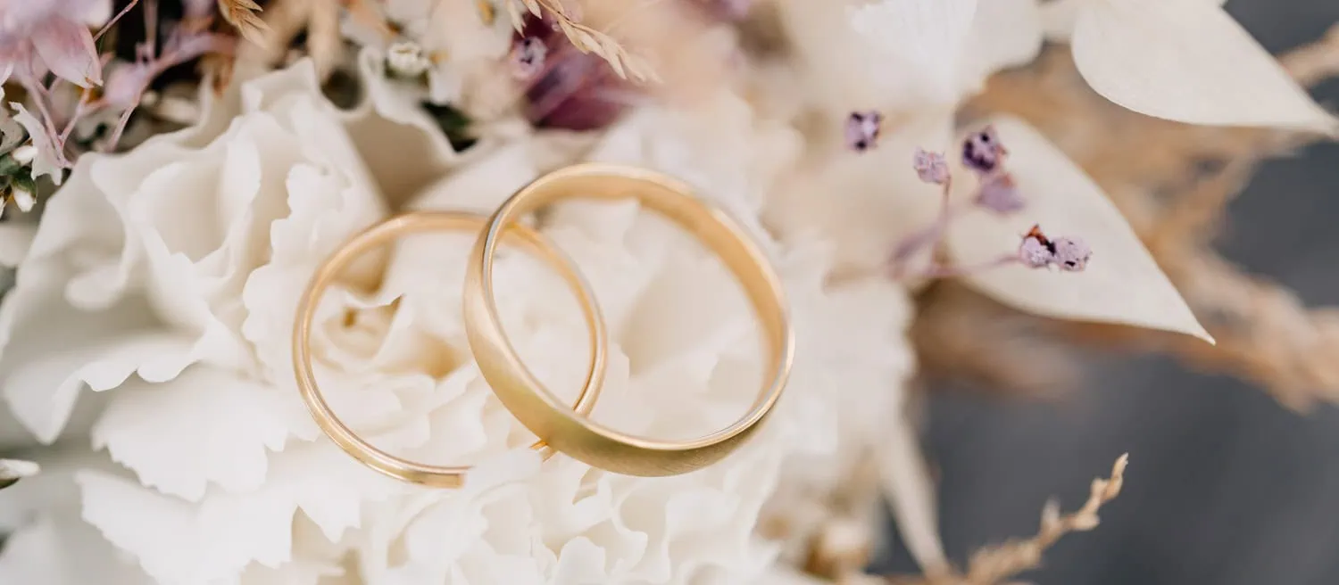 Das Bild zeigt zwei goldene Eheringe, die auf weißen Blumen liegen.