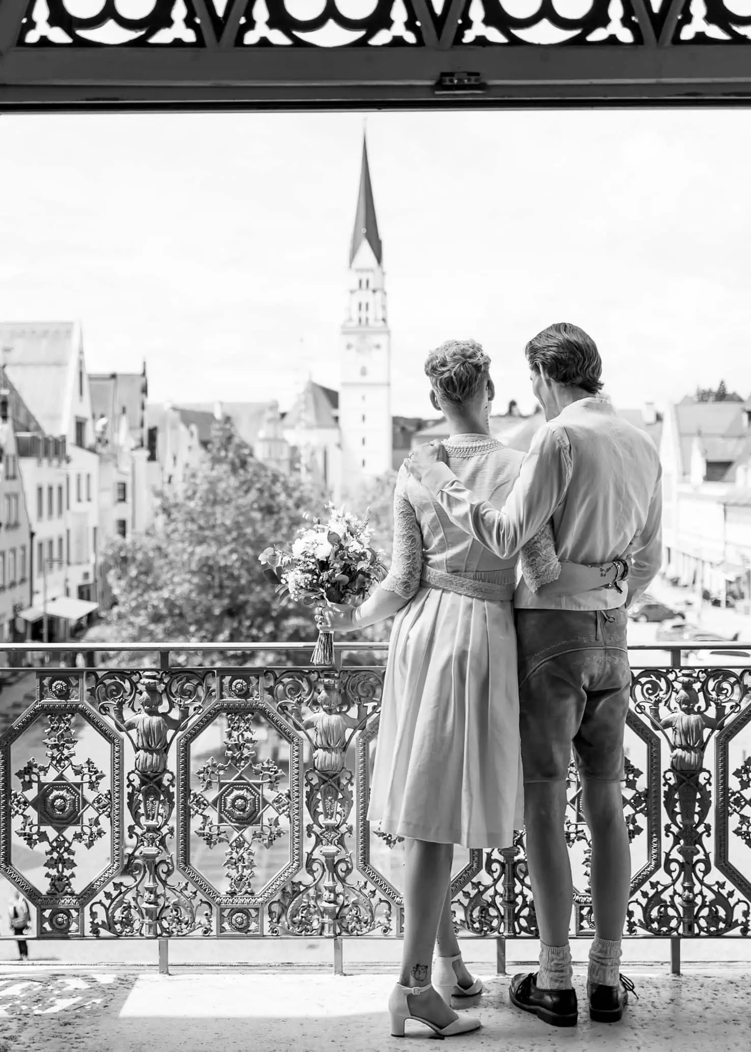 Zwei Personen, vermutlich ein Paar, stehen Arm in Arm auf einem Balkon und blicken auf eine Stadtlandschaft mit einem Kirchturm im Hintergrund.