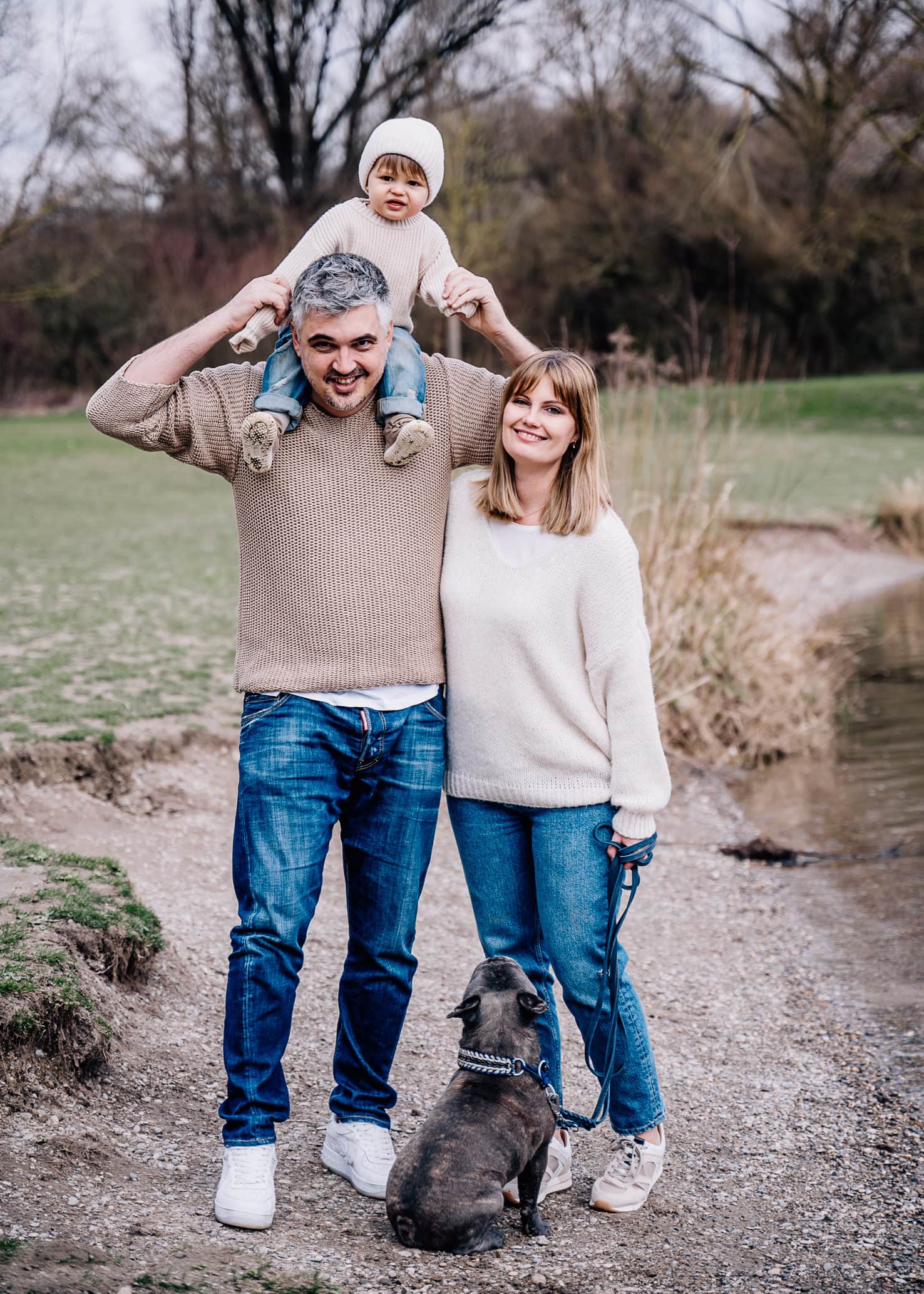 Das Bild zeigt eine Familie mit einem Kind auf den Schultern eines Mannes und einem Hund an der Seite einer Frau im Freien.