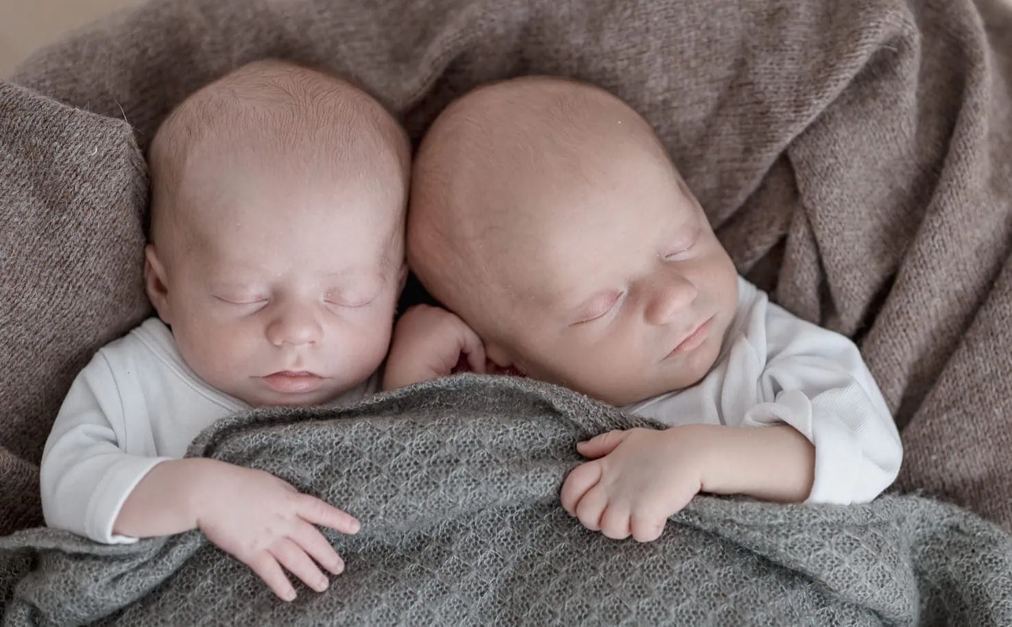 Das Bild zeigt zwei schlafende Babys, die nebeneinander liegen und mit einer grauen Decke zugedeckt sind.