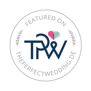 Das ist ein Logo mit der Aufschrift "Featured on ThePerfectWedding.de" umgeben von einem Lorbeerkranz und einem Herzmotiv.