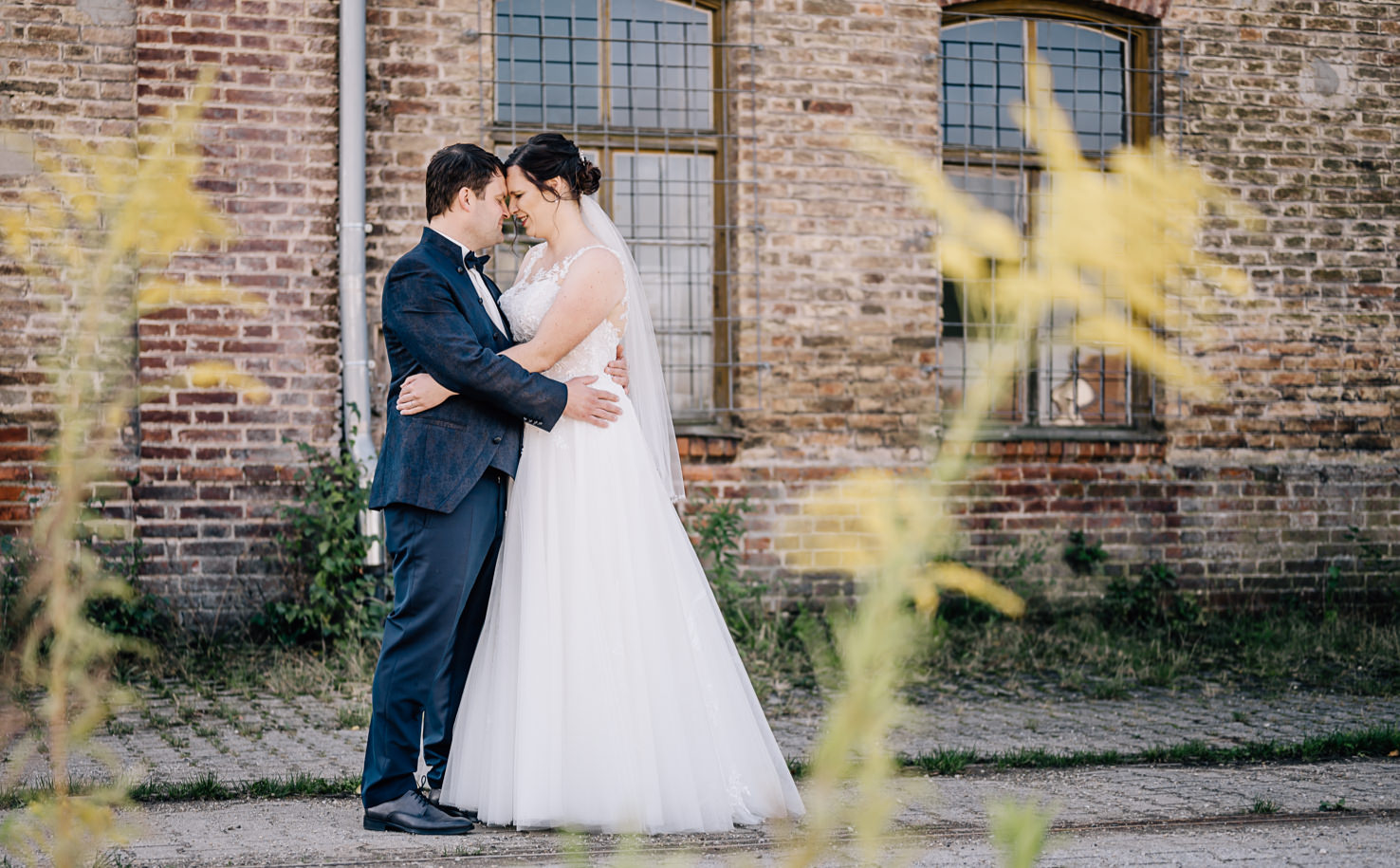 Zwei Personen in Hochzeitskleidung umarmen sich vor einer Ziegelmauer.