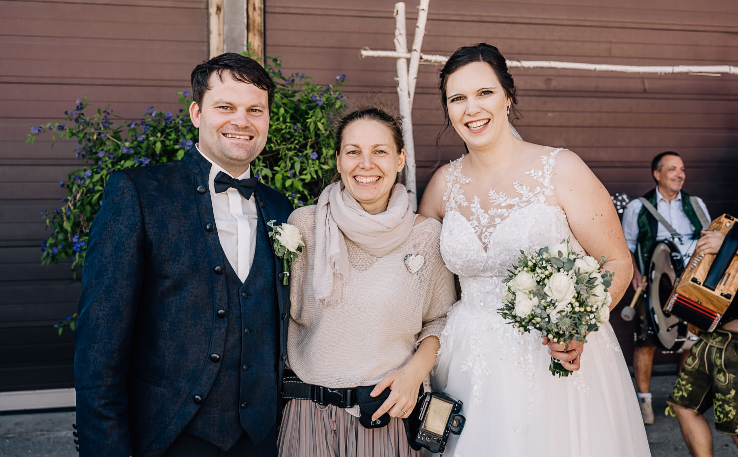 Das Bild zeigt zwei Personen in Hochzeitskleidung, die neben einer lächelnden Frau mit einer Kamera stehen, mit einem Musiker im Hintergrund.