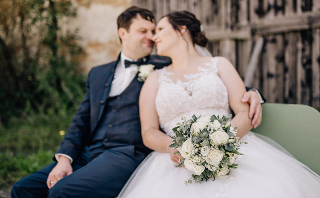 Das Bild zeigt ein Brautpaar in Hochzeitskleidung, das auf einer Bank sitzt und Zuneigung ausstrahlt.