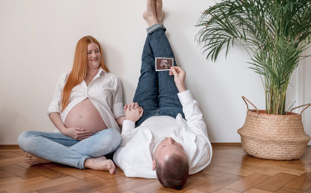 In diesem liebevollen Schwangerschaftsbild von Fotografin Mica Zeitz posiert ein Paar entspannt in einem modernen, hellen Raum, während sie voller Vorfreude auf ihr Babyblicken. Die Kombination aus der sanften Atmosphäre und dem Ultraschallbild symbolisiert die bevorstehende Elternschaft und die Verbundenheit zwischen Partnern während der aufregenden Reise der Schwangerschaft.