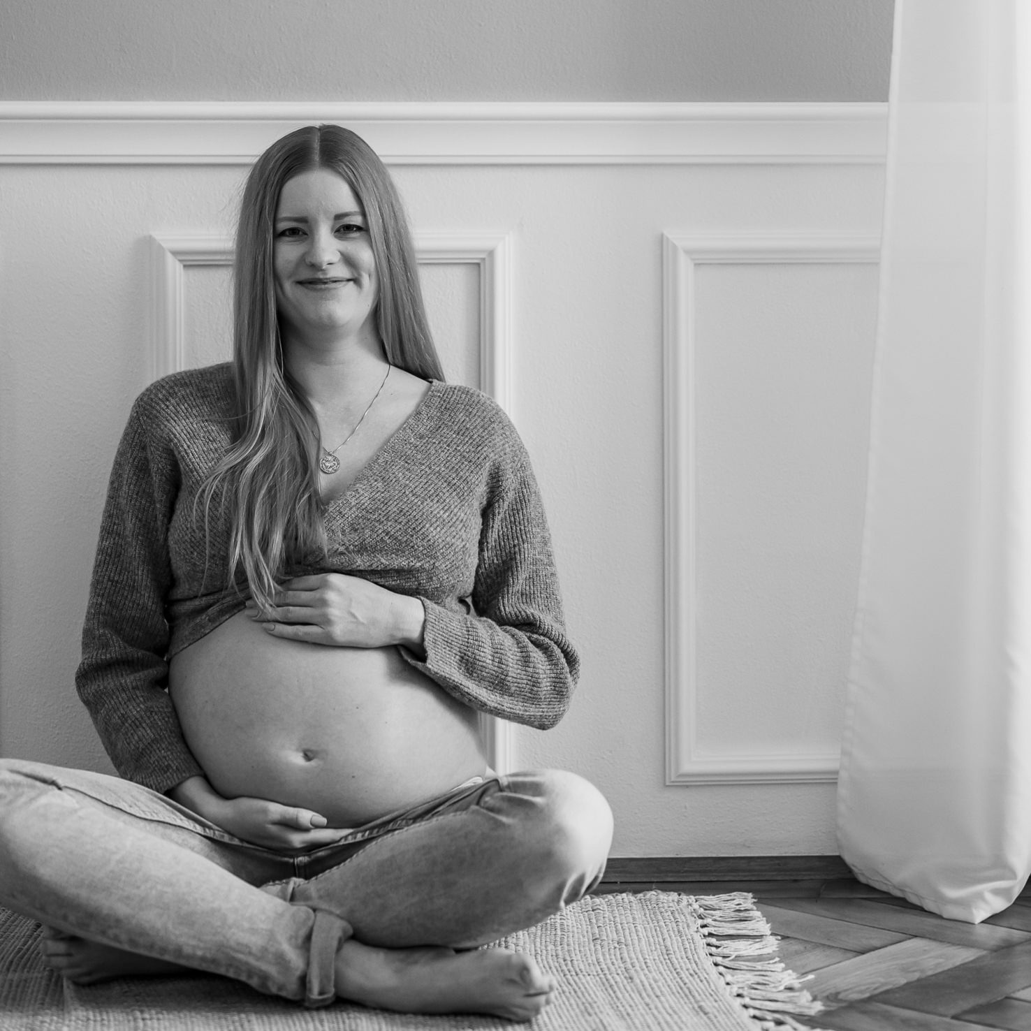 Das beeindruckende Schwarz-Weiß-Foto von Mica Zeitz zeigt eine werdende Mutter, die entspannt und glücklich auf einem Teppich sitzt und ihren Babybauch liebevoll umfasst. Der intime Moment verkörpert die Vorfreude auf das neue Leben und ist perfekt für die Themen Schwangerschaftsfotografie und Familienporträts.