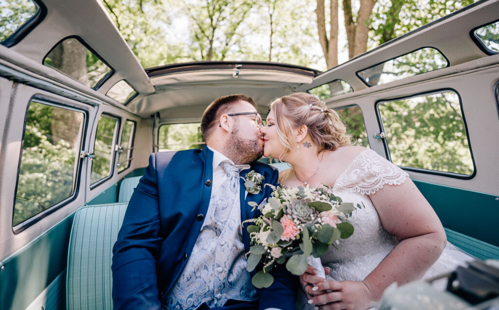 Dieses romantische Hochzeitsbild von Mica Zeitz zeigt ein glückliches Brautpaar, das sich innig küsst, während sie in einem nostalgischen VW-Bus sitzen. Die idyllische Kulisse und die liebevoll gestalteten Blumenarrangements unterstreichen die emotionale Stimmung dieses unvergesslichen Moments.