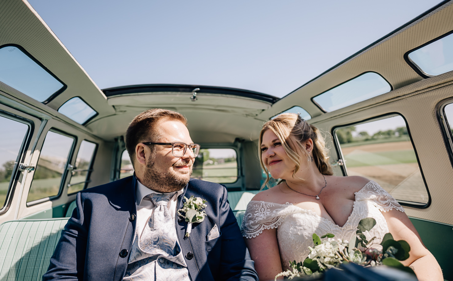 Das Bild zeigt ein Brautpaar, das in einem klassischen VW-Bus sitzt, während sie sich liebevoll anschauen und die strahlende Sonne genießen. Diese emotionale Momentaufnahme wurde von der talentierten Fotografin Mica Zeitz festgehalten und strahlt die Freude eines unvergesslichen Hochzeitstags aus.