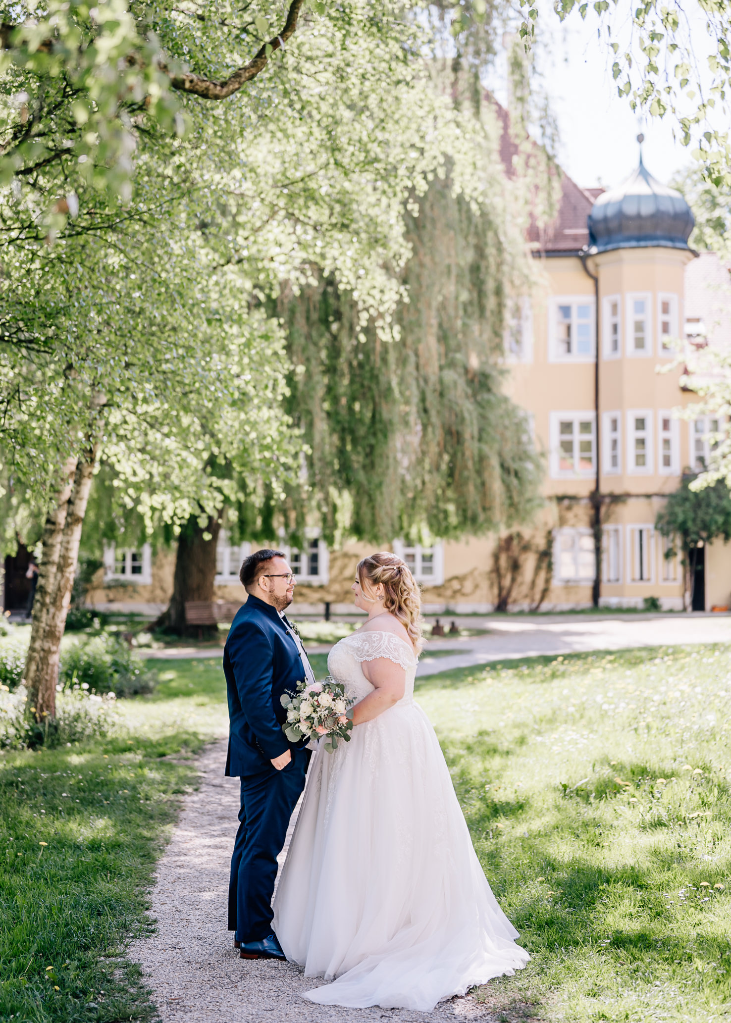 Dieses romantische Hochzeitsfoto von Mica Zeitz zeigt ein Paar, das sich in einem malerischen Garten umgeben von üppigem Grün und einem historischen Gebäude gegenübersteht. Die strahlende Freude und das liebevolle Band zwischen den Verliebten werden durch das sanfte Licht und die natürliche Kulisse perfekt eingefangen.