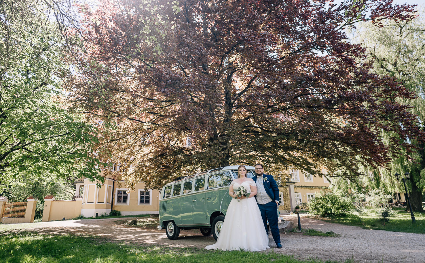 Das romantische Hochzeitsfoto von Mica Zeitz fängt das glückliche Paar vor einem nostalgischen VW-Bus und einem prächtigen Baum in voller Blüte ein, was die liebevolle Atmosphäre des besonderen Tages verstärkt. Diese bildhafte Darstellung verbindet natürliche Schönheit mit zeitlosem Hochzeitscharme und ist perfekt für Hochzeitsfotografie-Fans.