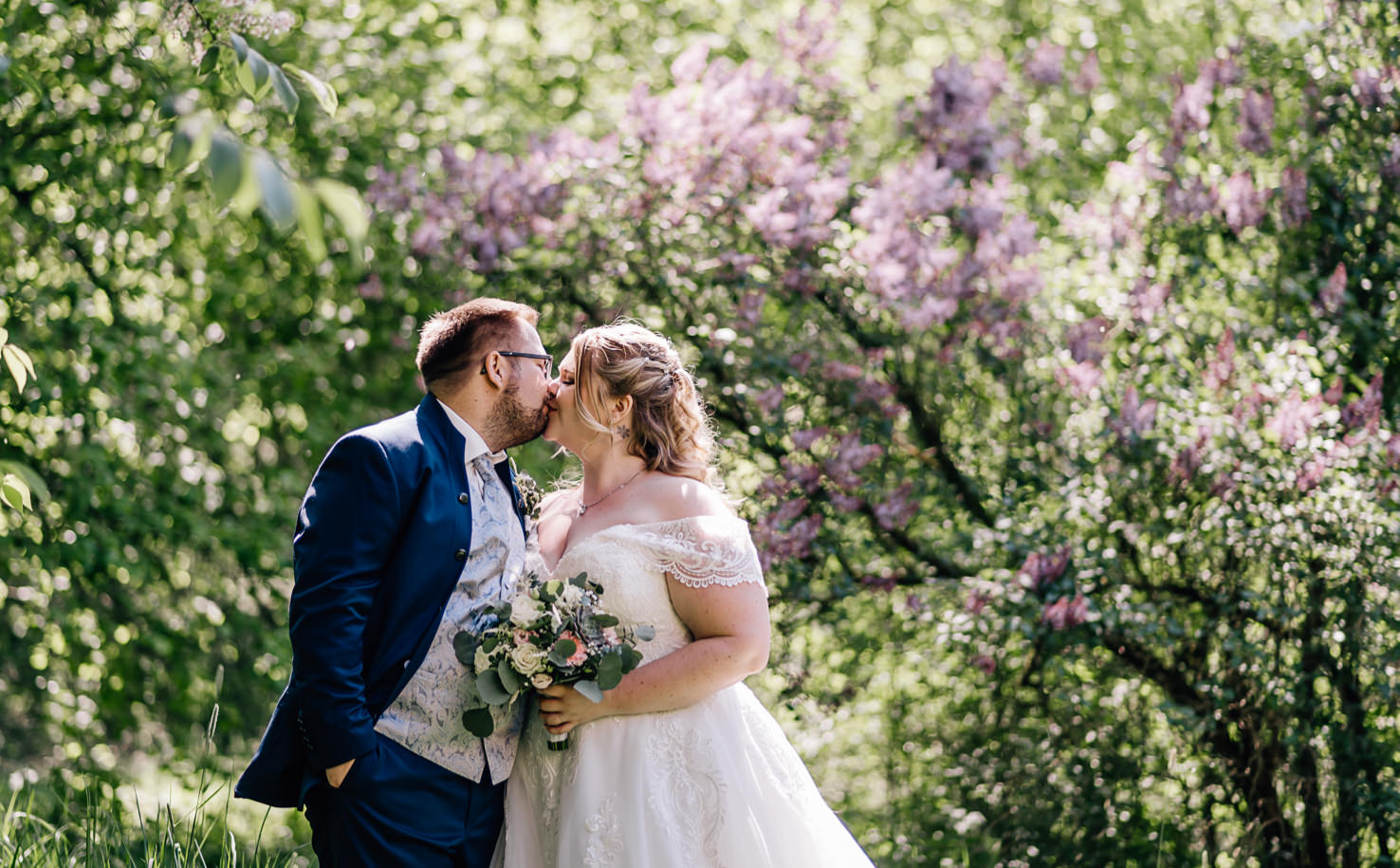 Im romantischen Grün eines blühenden Gartens zeigt die Fotografin Mica Zeitz ein verliebtes Paar, das sich sanft küsst und in festlicher Hochzeitskleidung erstrahlt. Die wundervolle Kulisse, umgeben von lilafarbenen Blüten, verleiht dieser Szene eine zauberhafte, unvergessliche Atmosphäre für Hochzeitsfotografie.