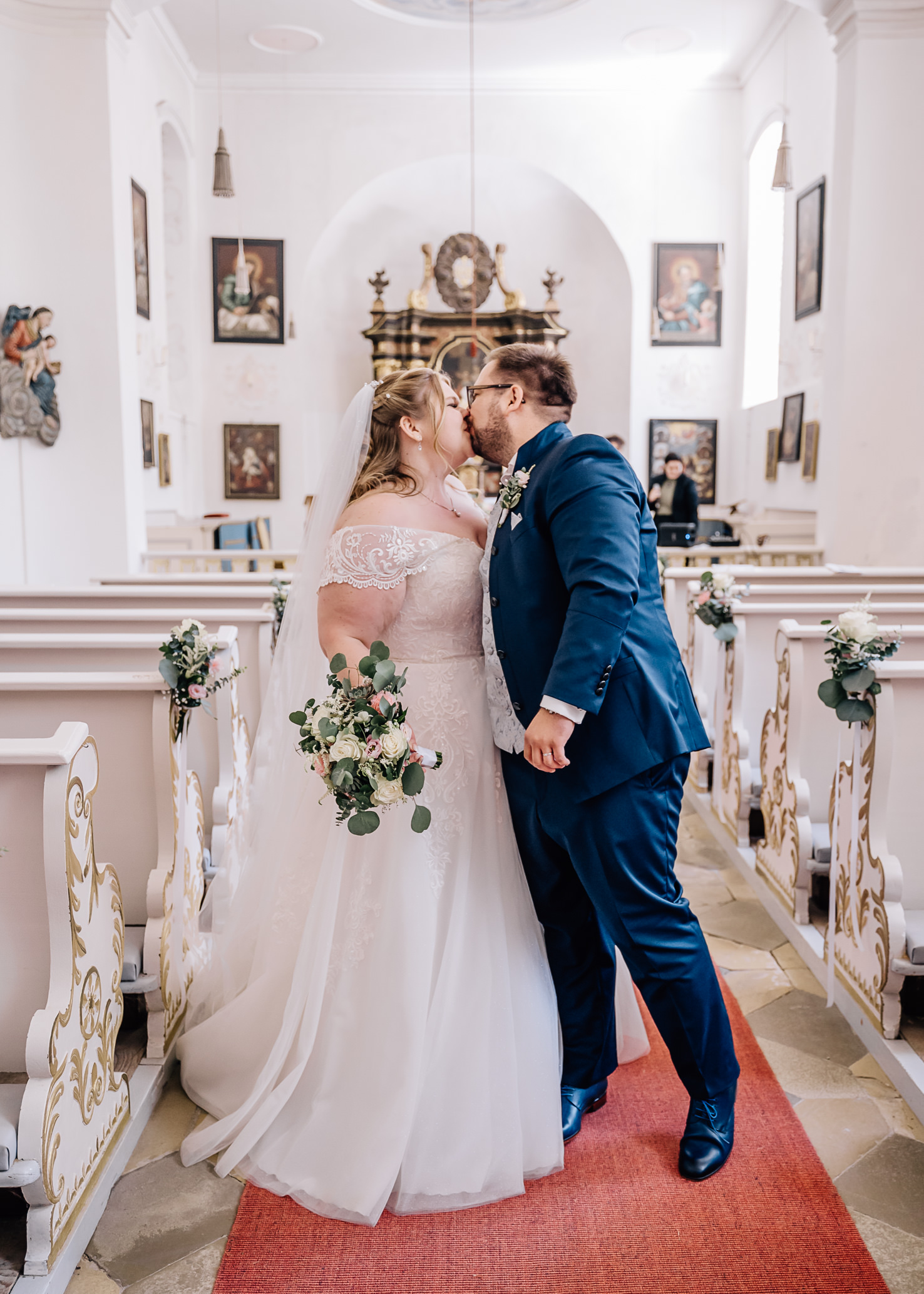 In dieser romantischen Hochzeitsaufnahme von Mica Zeitz küssen sich das Brautpaar in einer malerischen Kirche, umgeben von eleganten Kirchenbänken und kunstvollen Wandgemälden. Die Szene strahlt Liebe und Zweisamkeit aus und fängt den besonderen Moment ihres großen Tages perfekt ein.
