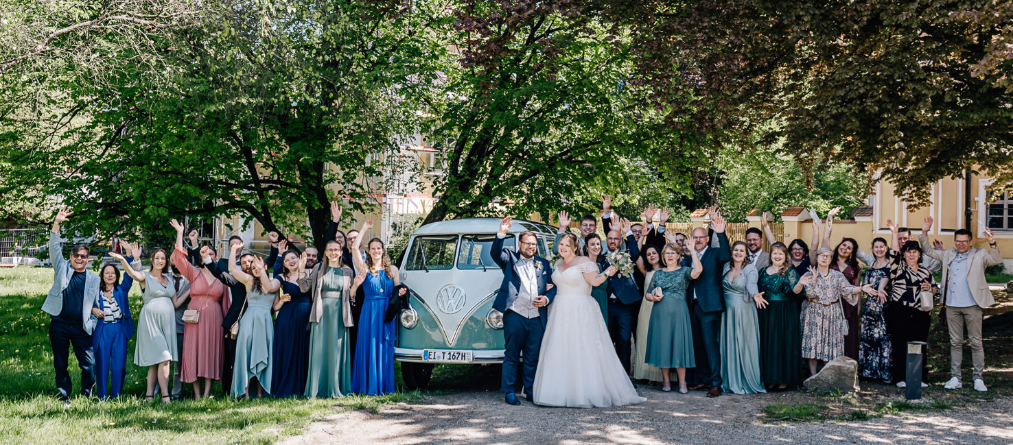 Das Bild zeigt eine fröhliche Hochzeitsgesellschaft in einem malerischen Garten, posierend vor einem nostalgischen VW-Bus, während sie ihre Freude mit erhobenen Händen ausdrücken. Diese stimmungsvolle Szene, festgehalten von der talentierten Fotografin Mica Zeitz, verkörpert den Zauber von unvergesslichen Hochzeitsmomenten und perfekt abgestimmten Hochzeitsoutfits.