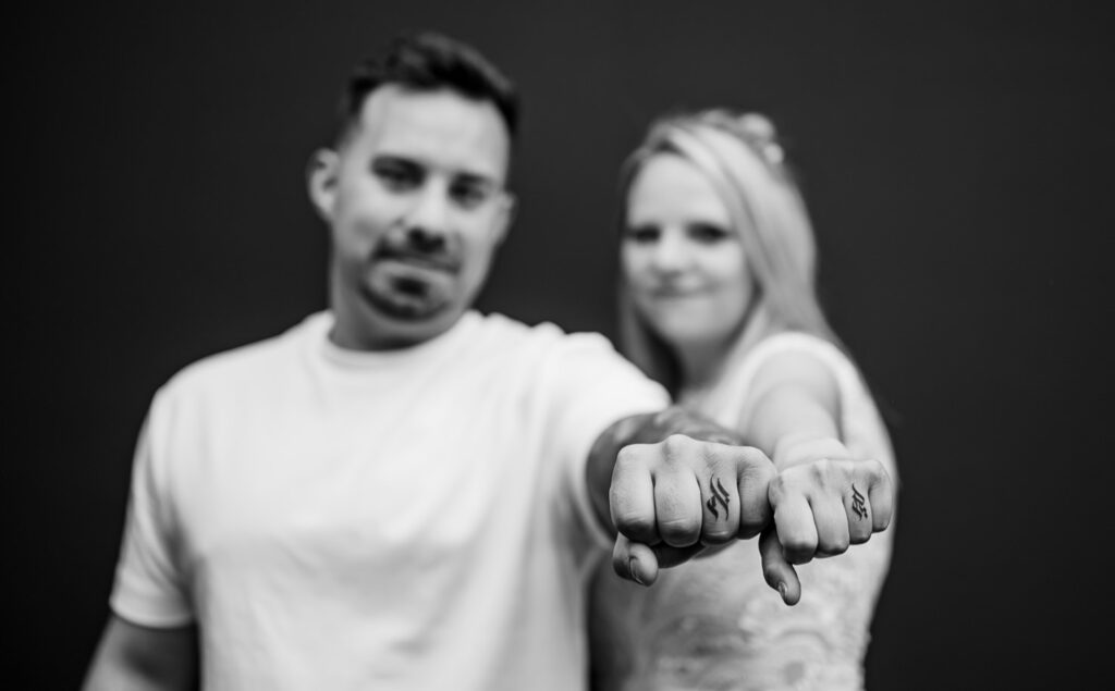 Dieses eindrucksvolle Porträt von Mica Zeitz zeigt ein Paar, das stolz seine symbolischen Tattoos präsentiert, was die tiefe Verbundenheit und die Emotionen zwischen ihnen unterstreicht. Die klare Fokussierung auf die Hände betont die Intimität und die persönlichen Geschichten, die hinter den Tattoos stehen.