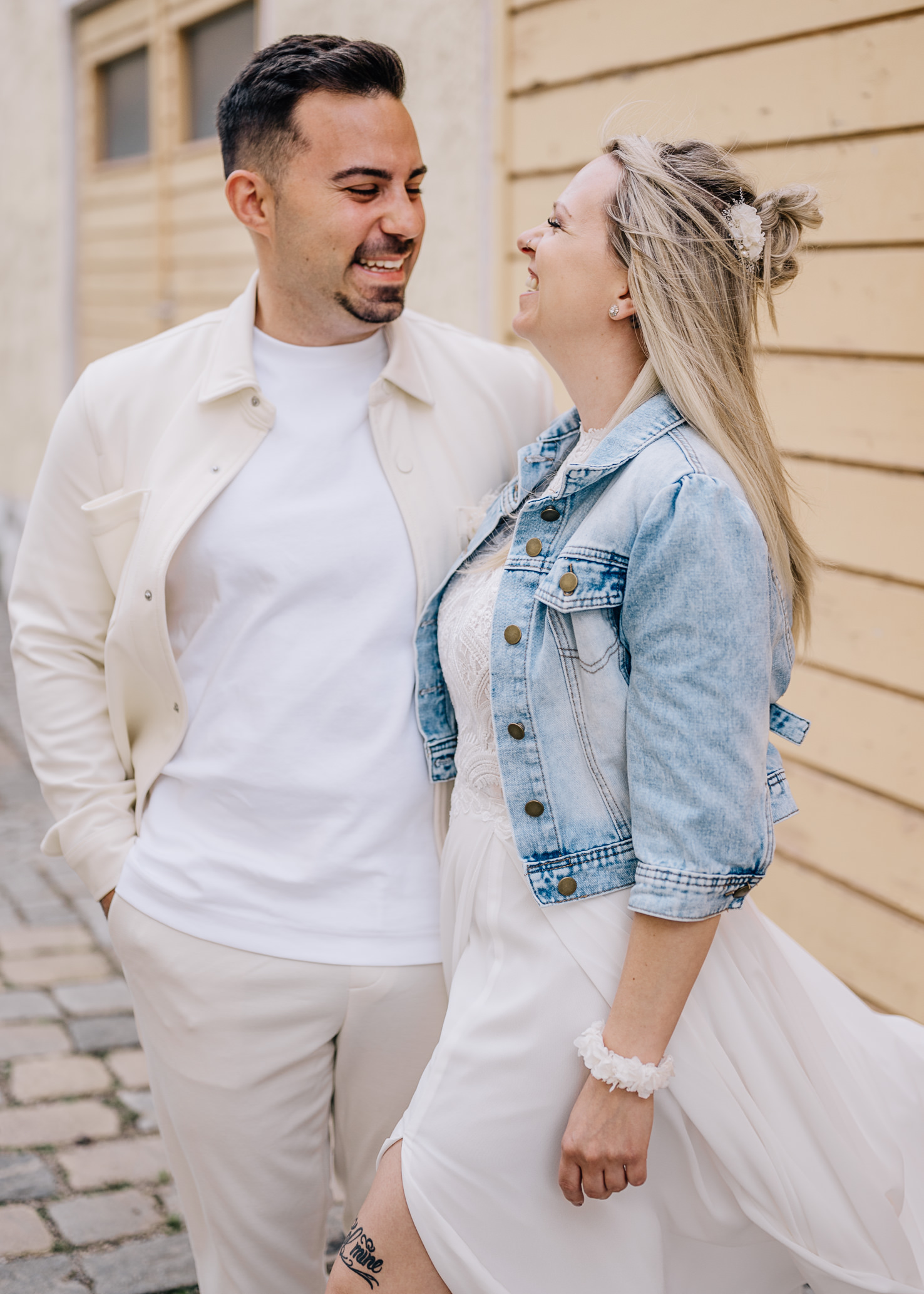 Das Bild von Mica Zeitz zeigt ein glückliches Paar in einem verträumten, urbanen Setting, in dem Liebe und Freude in der Luft liegen. Die stilvolle Kleidung, kombiniert mit einem lässigen Lächeln, spiegelt eine moderne, romantische Atmosphäre wider, ideal für Hochzeitsfotos oder Paarshootings.