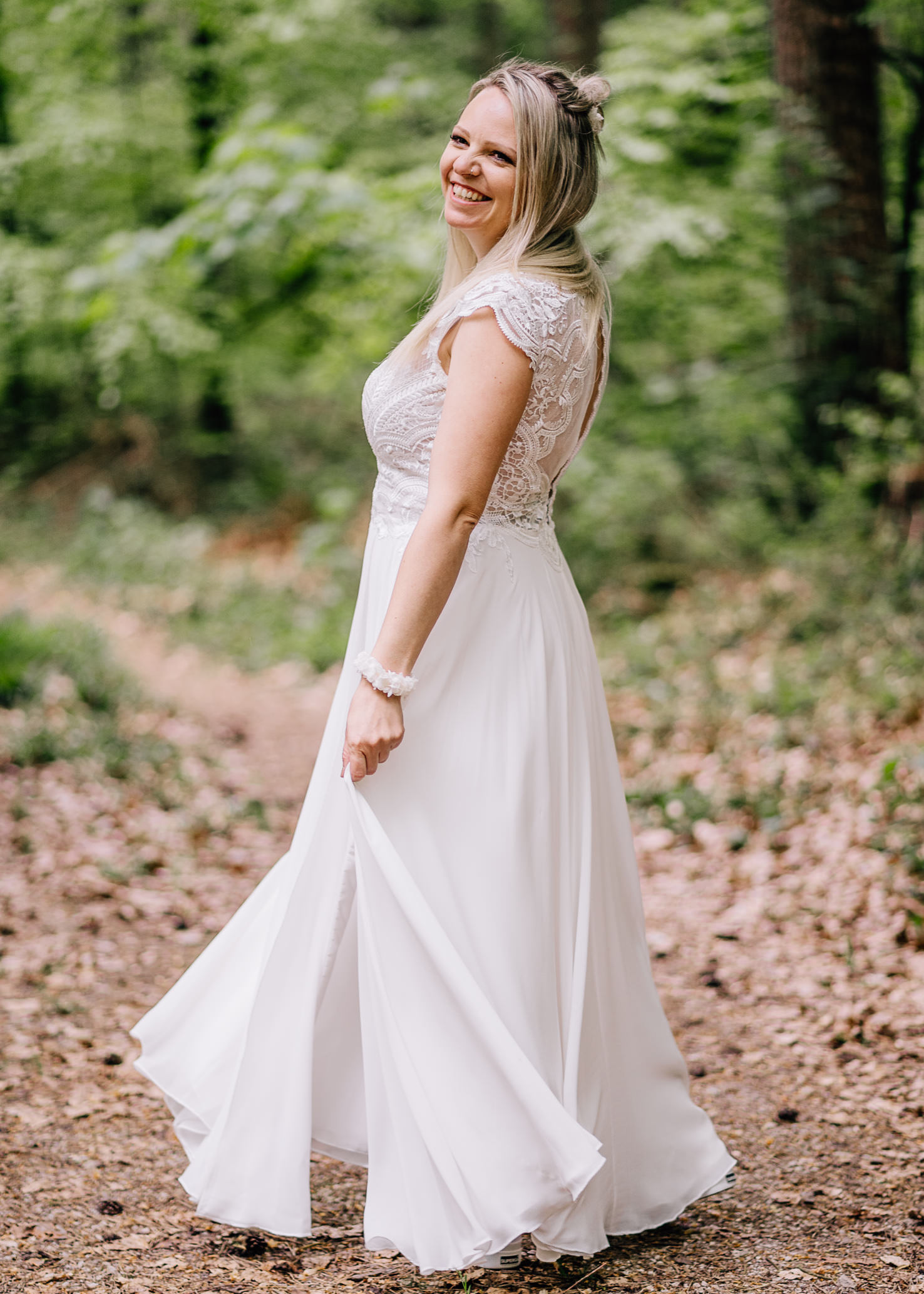 In einem malerischen Waldumfeld strahlt die Braut in einem eleganten, fließenden weißen Kleid von Fotografin Mica Zeitz. Ihr fröhliches Lächeln und die zarte Stilisierung verleihen dem Bild eine romantische und verträumte Atmosphäre, ideal für Hochzeitsfotografie.