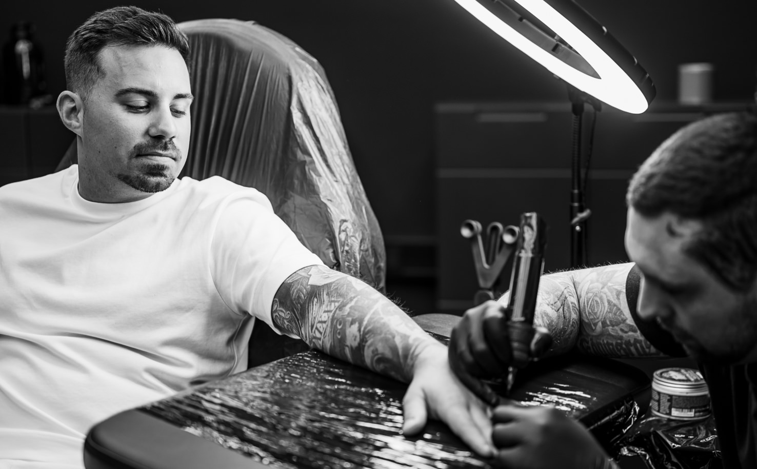 Das beeindruckende Schwarz-Weiß-Bild von Fotografin Mica Zeitz zeigt einen tätowierten Mann, der entspannt in einem Tattoo-Studio sitzt, während ein Künstler konzentriert an seinem Arm arbeitet. Die Aufnahme fängt die Intensität des kreativen Prozesses und die Kunstfertigkeit des Tätowierers perfekt ein und beleuchtet die wachsende Beliebtheit von Körperkunst.