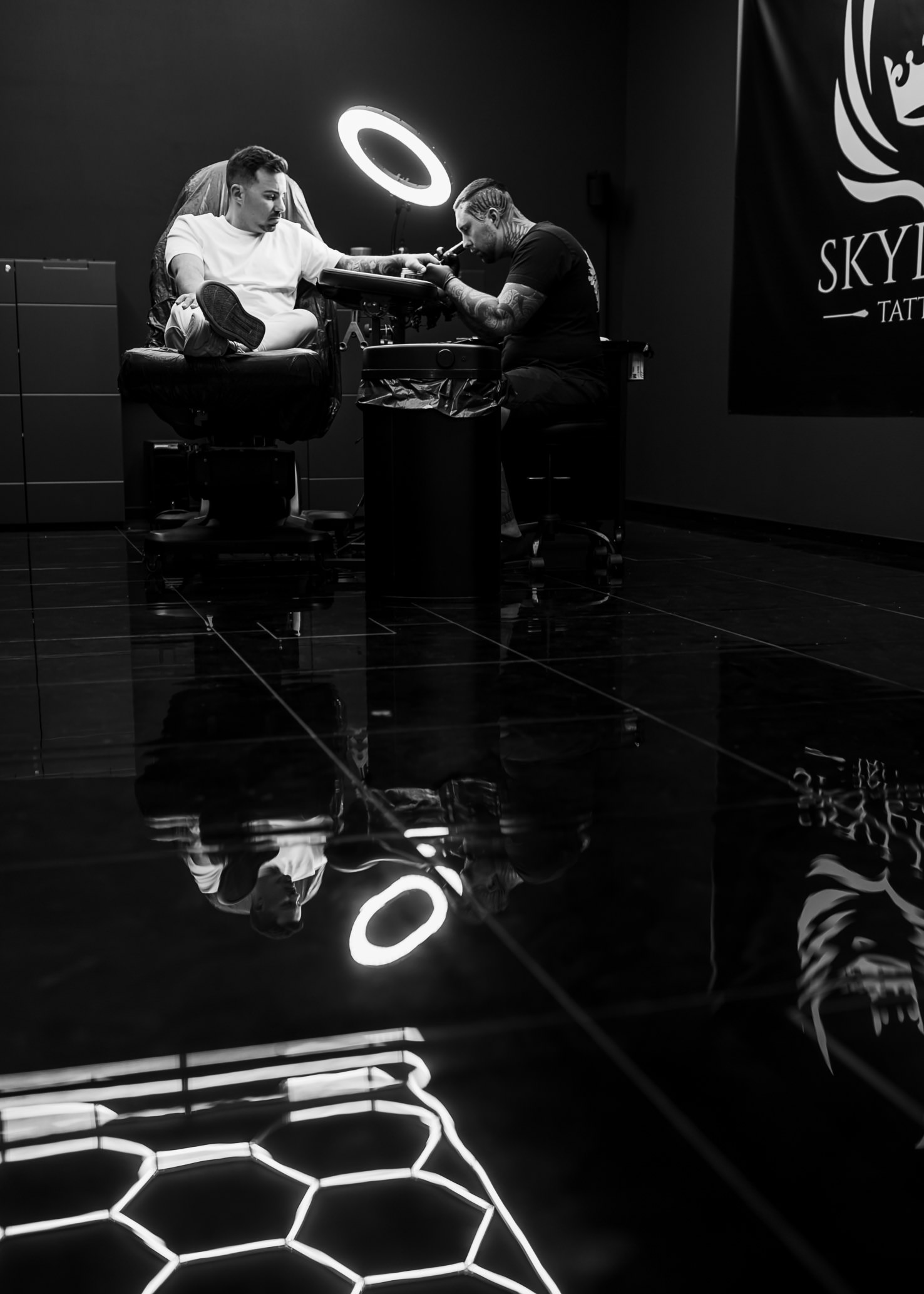 In diesem eindrucksvollen Schwarz-Weiß-Bild von Mica Zeitz wird ein Tattoo-Prozess in einem modernen Studio festgehalten, wobei die dramatische Beleuchtung und der Glanz des Bodens die intime Atmosphäre unterstreichen. Der Fokus auf den Künstler und den Klienten hebt die Kunst des Tätowierens und die Fähigkeiten des Tätowierers hervor, was für Liebhaber von Körperkunst und Tattoo-Designs von großem Interesse ist.