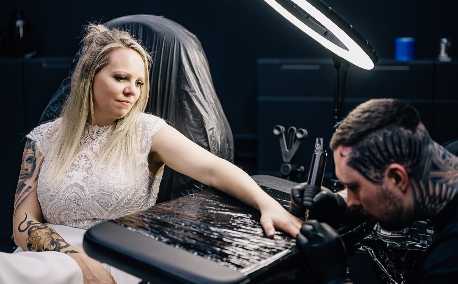 In diesem eindrucksvollen Bild von Fotografin Mica Zeitz wird der Moment eines Tattoos festgehalten, während eine junge Frau in einem elegantem, weißen Outfit in einem modernen Studio posiert. Das Zusammenspiel von Kunst und Körpermodifikation sowie die emotionale Intensität des Augenblicks verleihen dem Foto eine besondere Tiefe und Anziehungskraft.
