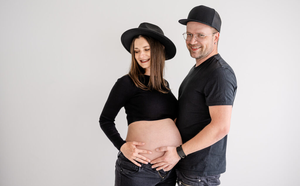 Dieses liebevolle Paar posiert in stilvollen schwarz-weißen Outfits, während sie die Vorfreude auf ihr kommendes Baby verkörpern. Fotografiert von Mica Zeitz, strahlt das Bild Wärme und Intimität aus und fängt einen besonderen Moment der Schwangerschaft perfekt ein.