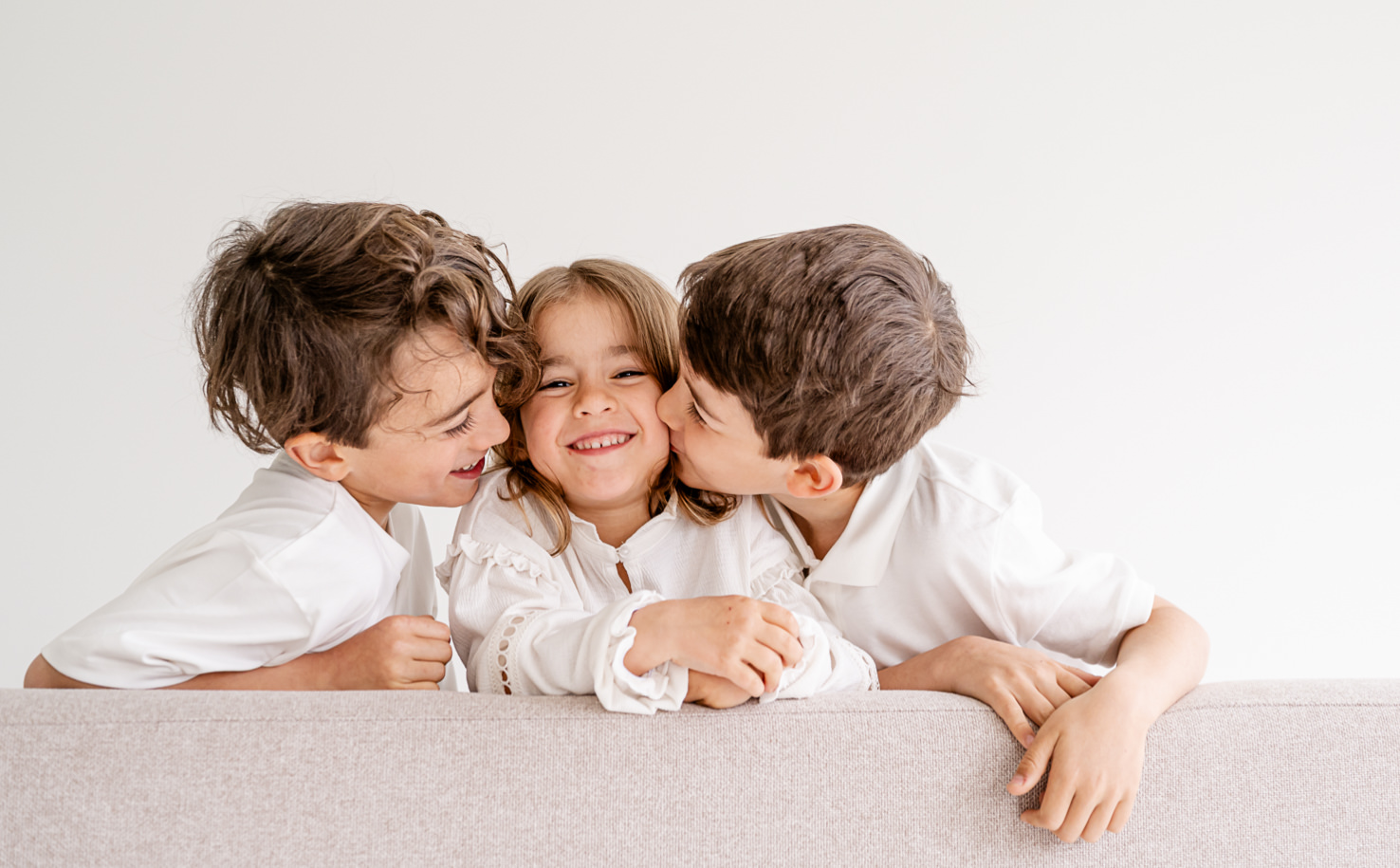 In diesem bezaubernden Familienporträt von Mica Zeitz strahlen Geschwisterliebe und kindliche Freude, während ein Mädchen von ihren Brüdern umarmt und geküsst wird. Die hellen Farben und die minimalistischen Hintergründe verleihen dem Bild eine einladende und fröhliche Atmosphäre.