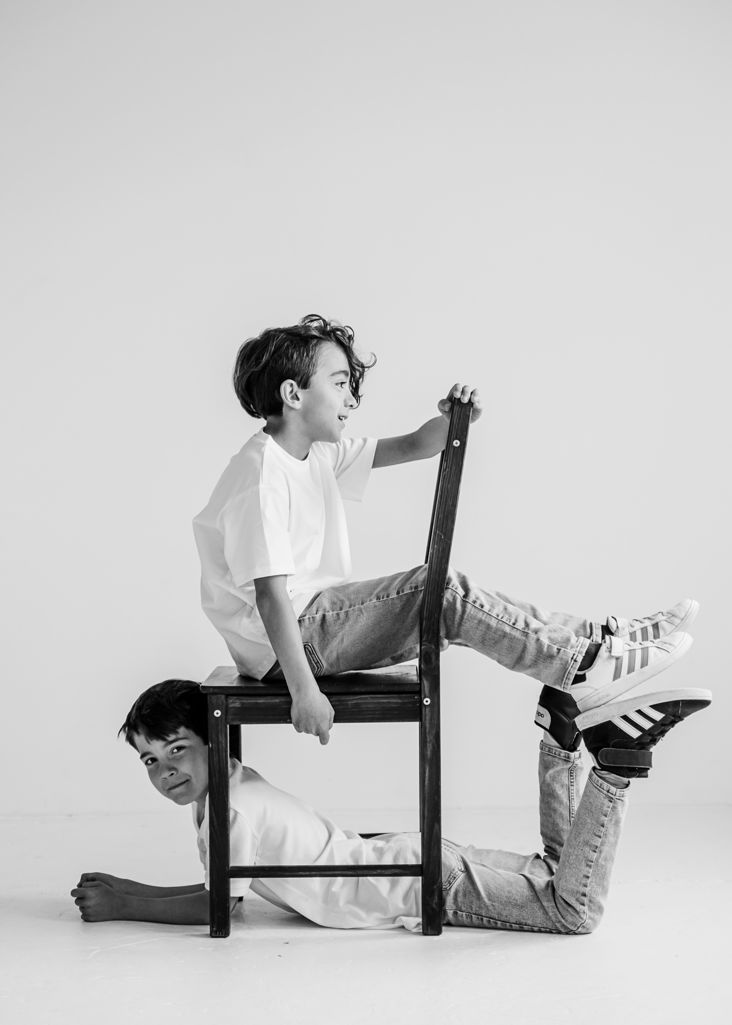 Das kreative Foto von Mica Zeitz zeigt zwei Jungen, die in einer spielerischen Pose auf einem Stuhl sitzen, was Dynamik und kindliche Unbeschwertheit ausstrahlt. Ein minimalistischer Hintergrund betont die lebhaften Interaktionen und das Geschick der Kinder, während die Schwarz-Weiß-Darstellung zeitlose Eleganz verleiht.