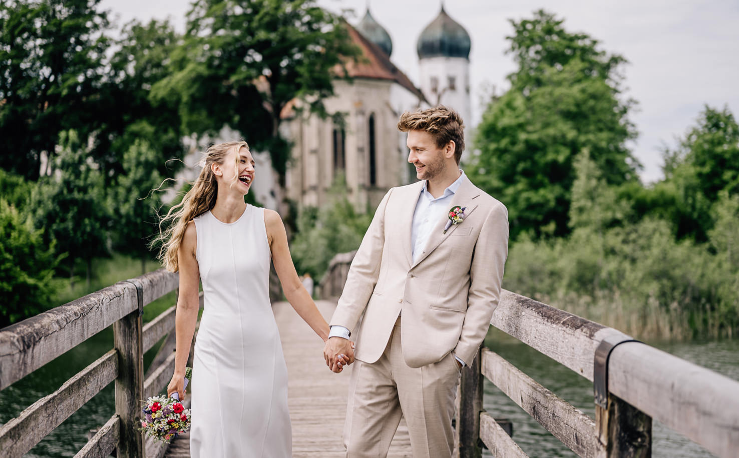 In dieser romantischen Hochzeitsaufnahme von Mica Zeitz strahlt das frisch vermählte Paar auf einer malerischen Holzbrücke, umgeben von üppigem Grün und historischer Architektur. Die lebendige Emotion und das Lachen der beiden fangen die Freude des Tages ein und machen dieses Bild zu einem perfekten Beispiel für Hochzeitsfotografie.