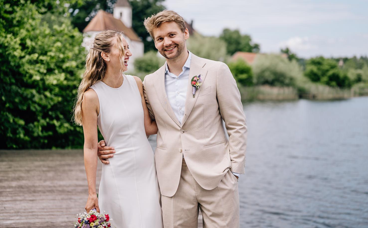Das Bild von der Fotografin Mica Zeitz fängt das glückliche Brautpaar in einer romantischen Umgebung am Wasser ein und strahlt Freude und Zuneigung aus. Mit den lebendigen Farben des Hochzeitsstraußes und der malerischen Kulisse wird der perfekte Moment für eine unvergessliche Hochzeitsfeier festgehalten.
