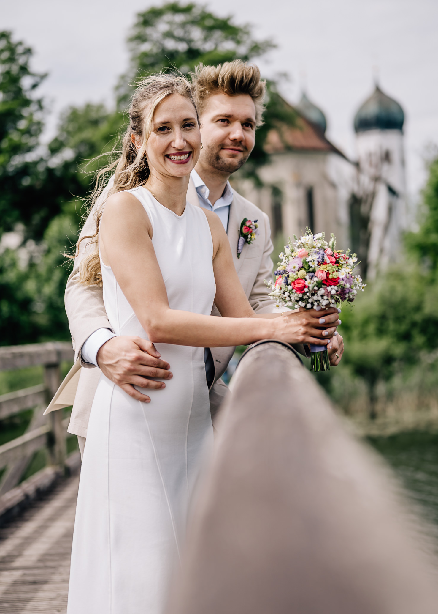 Auf diesem romantischen Hochzeitsfoto von Mica Zeitz strahlt das Brautpaar auf einer idyllischen Brücke, umgeben von üppigem Grün. Die wunderschöne Braut trägt ein elegantes, weißes Kleid und hält einen bunten Blumenstrauß, während der Bräutigam sie zärtlich umarmt, was die Liebe und Verbundenheit zwischen den beiden perfekt einfängt.