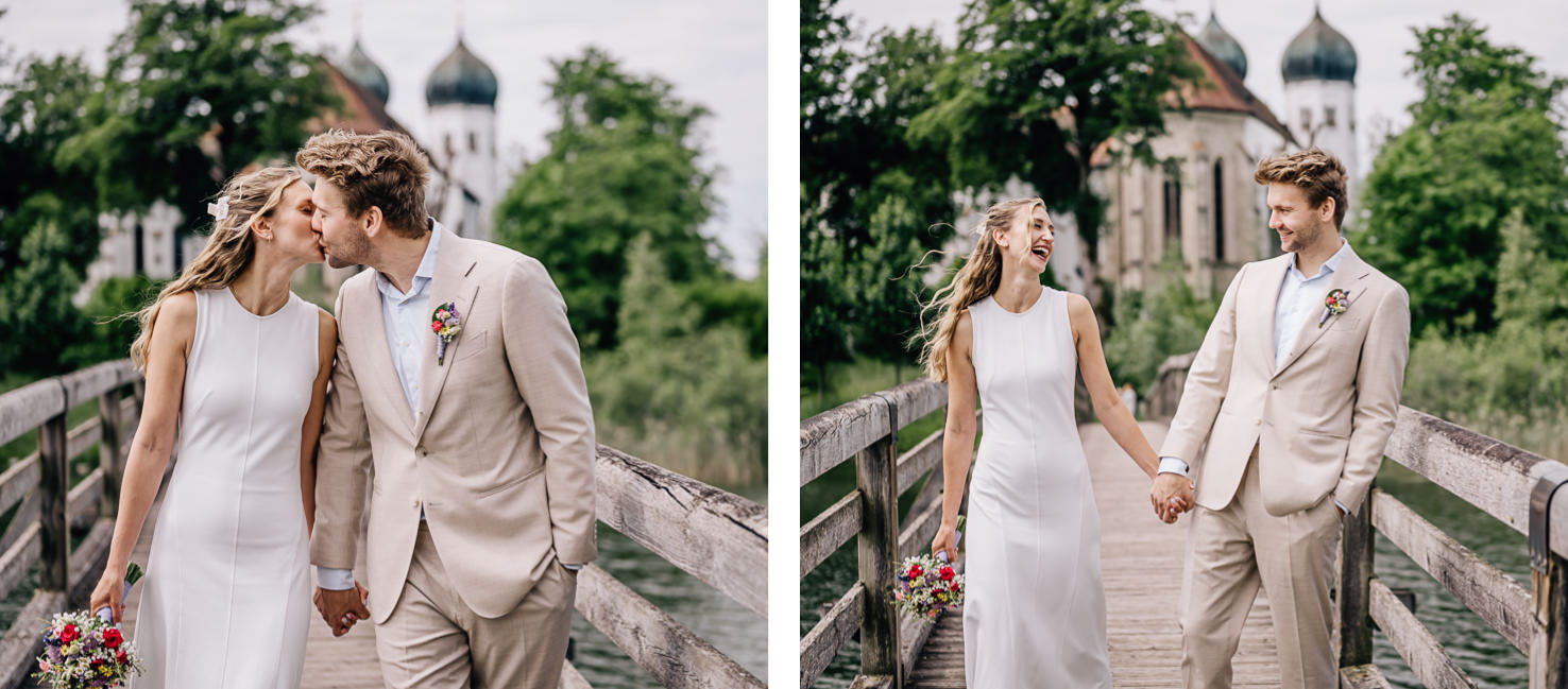 Ein romantisches Hochzeitsfoto von Mica Zeitz zeigt ein glückliches Paar, das Hand in Hand auf einer malerischen Brücke steht, umgeben von üppigem Grün und einem traumhaften Kirchenhintergrund. Die festlichen Outfits und die liebevollen Gesten strahlen Freude und Zuneigung aus, perfekt für Hochzeitsfotografie und Trauungserinnerungen.