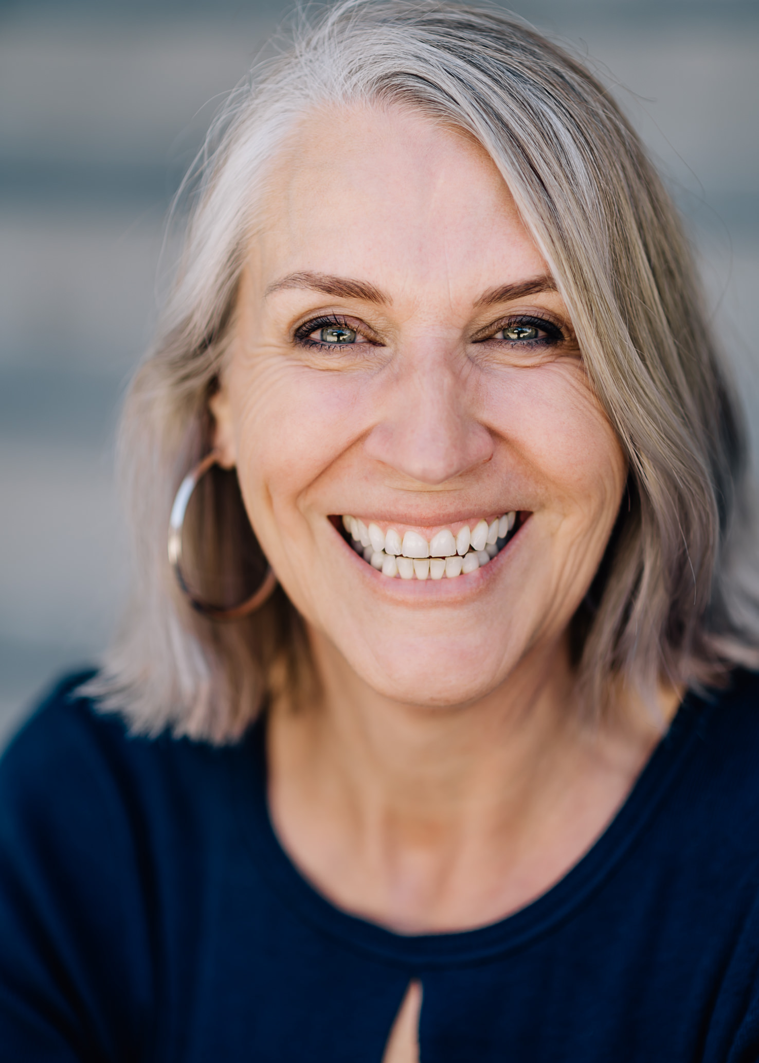Das fröhliche Porträt einer älteren Frau mit grauen Haaren, die in die Kamera lächelt, fängt die positive Ausstrahlung und Lebensfreude perfekt ein. Fotografiert von Mica Zeitz, strahlt dieses Bild Wärme und Authentizität aus, ideal für Themen wie Schönheit im Alter und natürliche Portraitfotografie.