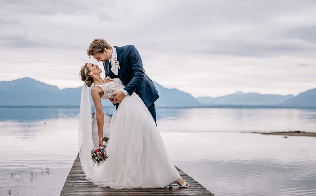 Das romantische Hochzeitsfoto von Mica Zeitz zeigt ein Brautpaar inmitten einer malerischen Seelandschaft, umrahmt von sanften Hügeln und dramatischem Himmel. Die emotionale Verbindung und der elegante Brautkleid-Look fangen die Essenz eines unvergesslichen Hochzeitstags ein.