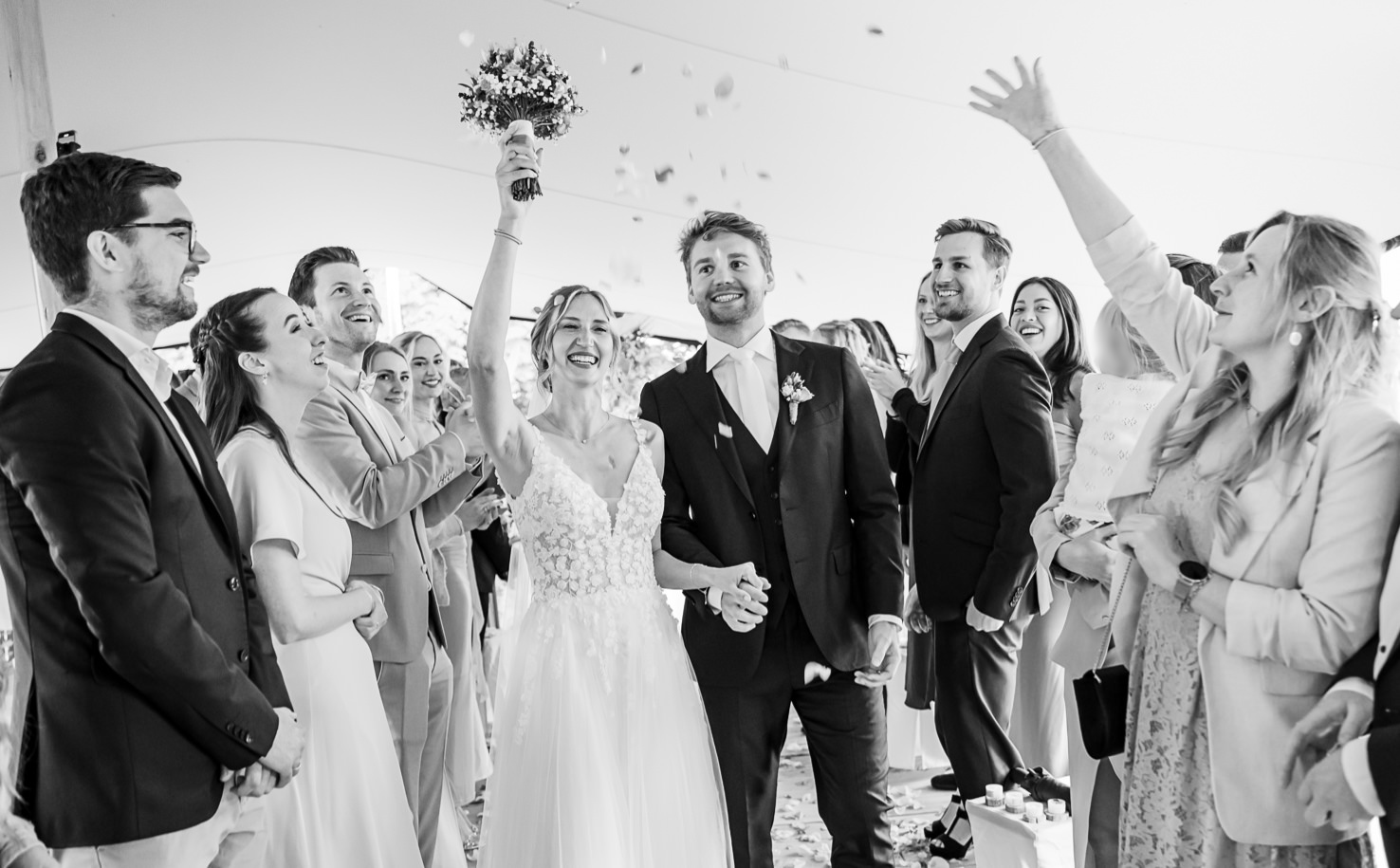 Auf diesem emotionalen Hochzeitsfoto von Mica Zeitz sehen wir das strahlende Brautpaar, das glücklich die Feierlichkeiten mit Freunden und Familie genießt, während Blütenblätter durch die Luft fliegen. Die Aufregung und die Freude des besonderen Moments machen dieses Bild zu einem perfekten Beispiel für unvergessliche Hochzeitsfotografie.