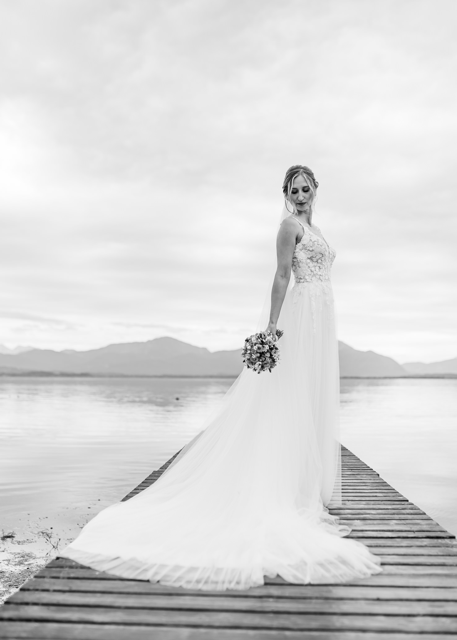 In diesem romantischen Hochzeitsporträt von Mica Zeitz strahlt die Braut in einem eleganten, fließenden Kleid auf einem Holzsteg, umgeben von der friedlichen Kulisse eines Sees und Bergen. Das schwarz-weiße Bild vermittelt eine zeitlose Eleganz, die jede Hochzeit unvergesslich macht.